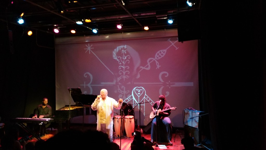 Durante uma apresentação musical do cantor James Germain, imagens relacionadas ao Voudou são projetadas na parede.